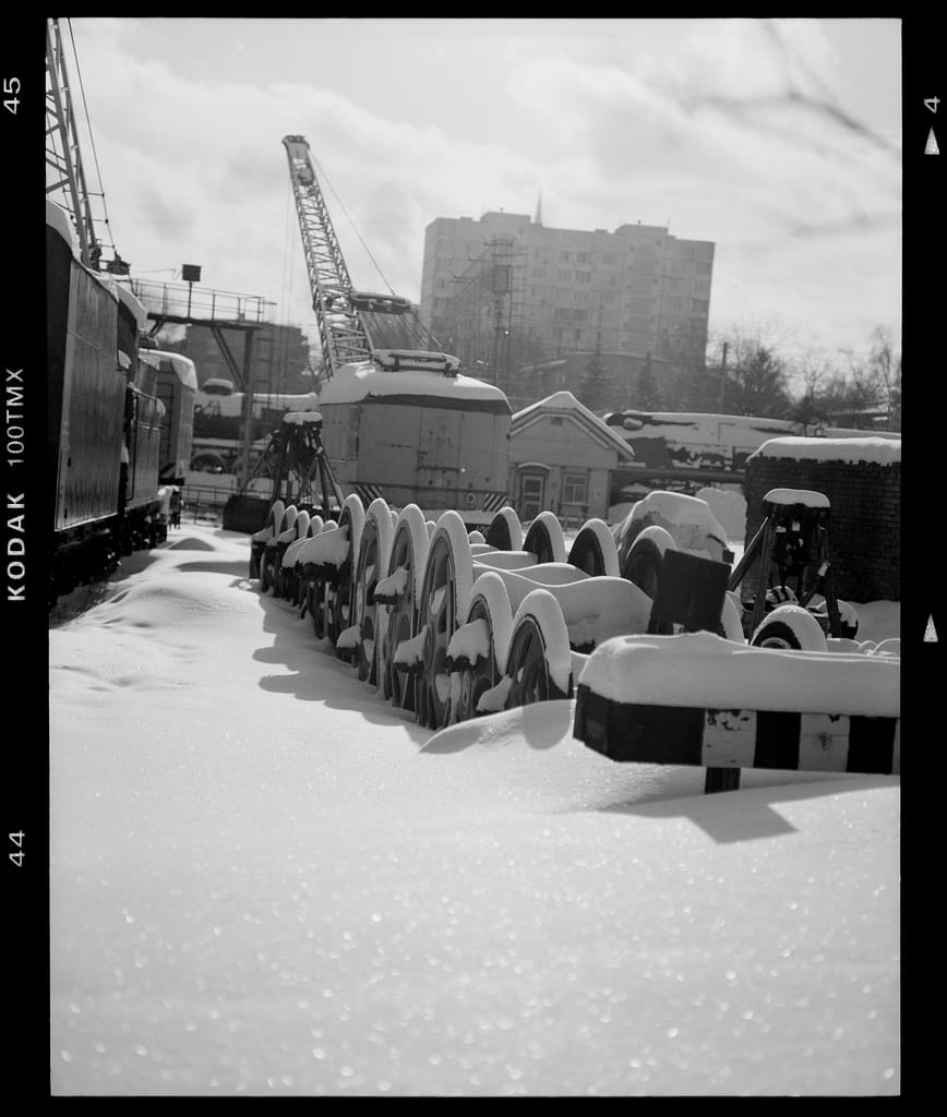 Black and white image of a snow-covered industrial yard with machinery and train parts.